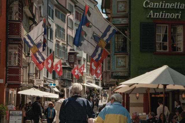 Zwei ältere Personen gehen Hand in Hand durch die Augustinergasse in Zürich, flankiert von historischen Häusern mit bunten Fassaden und Schweizer Fahnen. Vor den Restaurants sitzen Gäste an Tischen unter Sonnenschirmen, während die Straße lebendig wirkt
