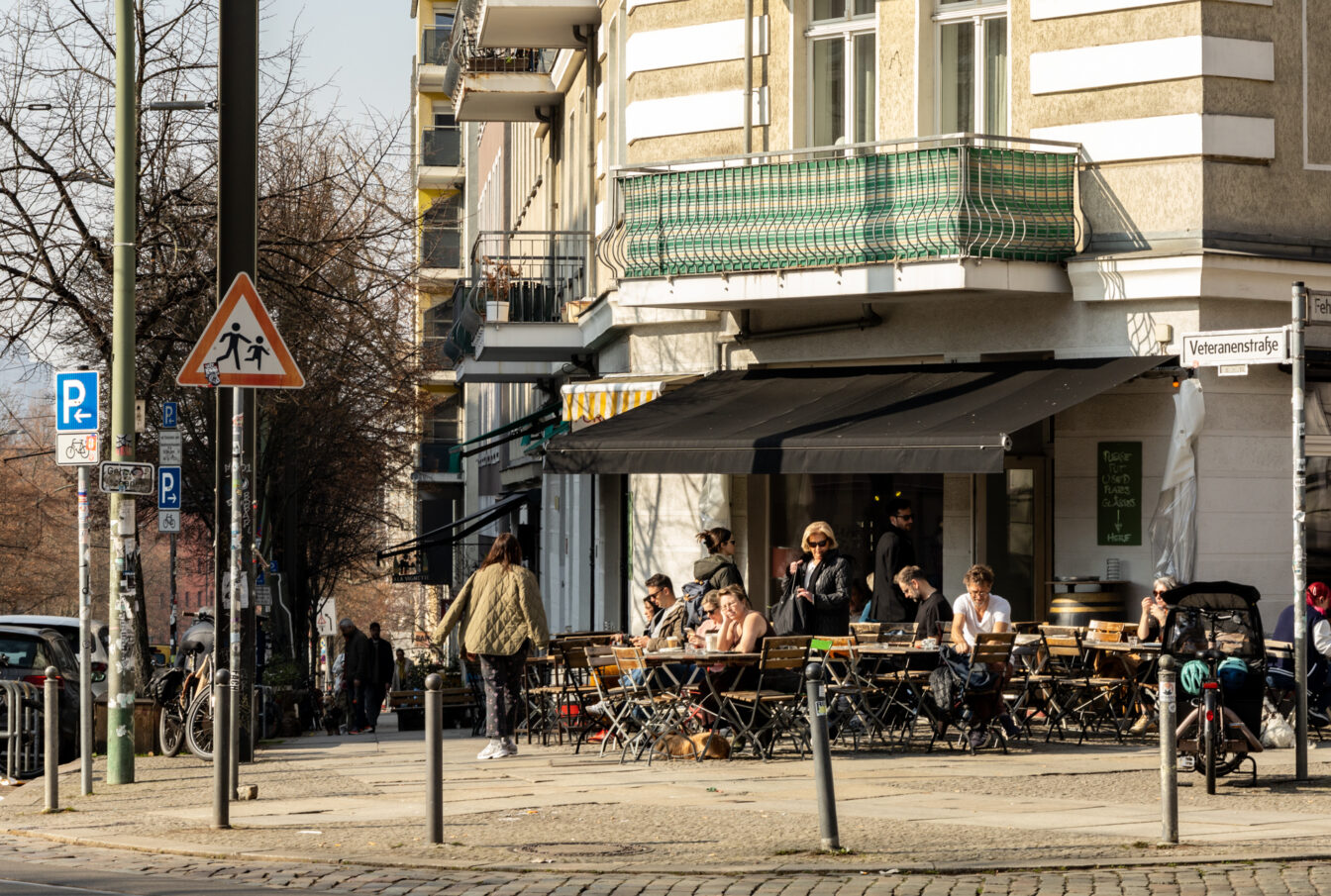 Ein typischer Berliner Straßenzug mit einer einladenden Café- und Bar-Szene. Die Szene spielt sich an der Veteranenstraße ab, wo Menschen das sonnige Wetter genießen und sich an den Tischen eines Straßencafés niederlassen. Schwarze Markisen spenden Schatten, während die Gäste in entspannter Atmosphäre sitzen, plaudern und ihr Essen oder Getränke genießen.