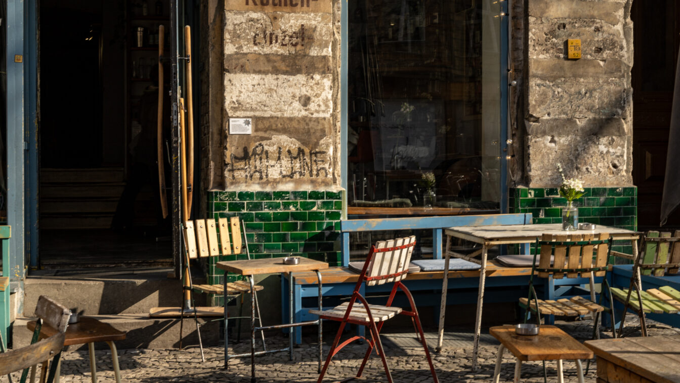 Ein charmantes Berliner Café mit einer rustikalen Fassade aus verwittertem Putz und grünen Fliesen. Vor dem Eingang stehen mehrere alte, teils wackelige Holztische und Stühle, darunter ein auffälliger weiß-roter Klappstuhl. Die Fenster spiegeln die Umgebung wider, und auf den Tischen stehen kleine Vasen mit weißen Blumen. Die Sonne wirft warme Lichtstrahlen und erzeugt lange Schatten auf dem Kopfsteinpflaster, während im Hintergrund eine geöffnete Tür den Blick ins Innere des Cafés freigibt.