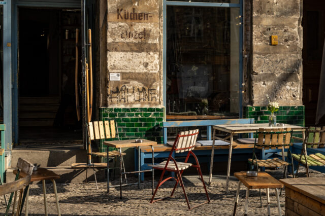Ein charmantes Berliner Café mit einer rustikalen Fassade aus verwittertem Putz und grünen Fliesen. Vor dem Eingang stehen mehrere alte, teils wackelige Holztische und Stühle, darunter ein auffälliger weiß-roter Klappstuhl. Die Fenster spiegeln die Umgebung wider, und auf den Tischen stehen kleine Vasen mit weißen Blumen. Die Sonne wirft warme Lichtstrahlen und erzeugt lange Schatten auf dem Kopfsteinpflaster, während im Hintergrund eine geöffnete Tür den Blick ins Innere des Cafés freigibt.