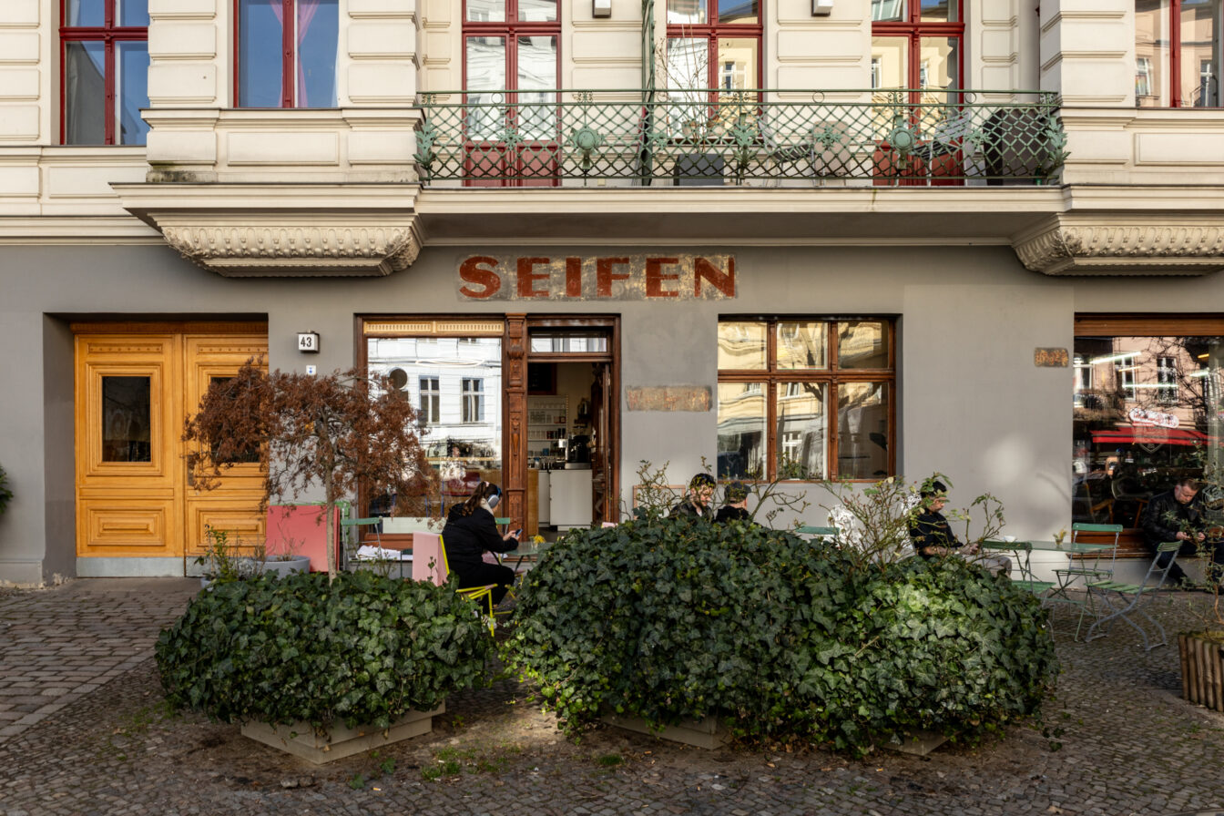 Ein charmantes Café in einem historischen Gebäude in der Oderberger Straße in Berlin. Über dem Eingang ist der verblasste Schriftzug 'SEIFEN' zu sehen, ein Hinweis auf die frühere Nutzung des Ladens. Die graue Fassade mit großen Fenstern und einer verzierten weißen Etage darüber verleiht dem Gebäude eine nostalgische Atmosphäre. Vor dem Café sitzen Menschen an bunten Metallstühlen und genießen das milde Wetter. Im Vordergrund sind grüne Pflanzen und ein kleiner, vertrockneter Baum in einem Pflanzkübel zu sehen. Das Café-Interieur ist durch die offene Tür und die Fenster sichtbar, während sich das Stadtleben in den Spiegelungen der Fensterscheiben widerspiegelt.