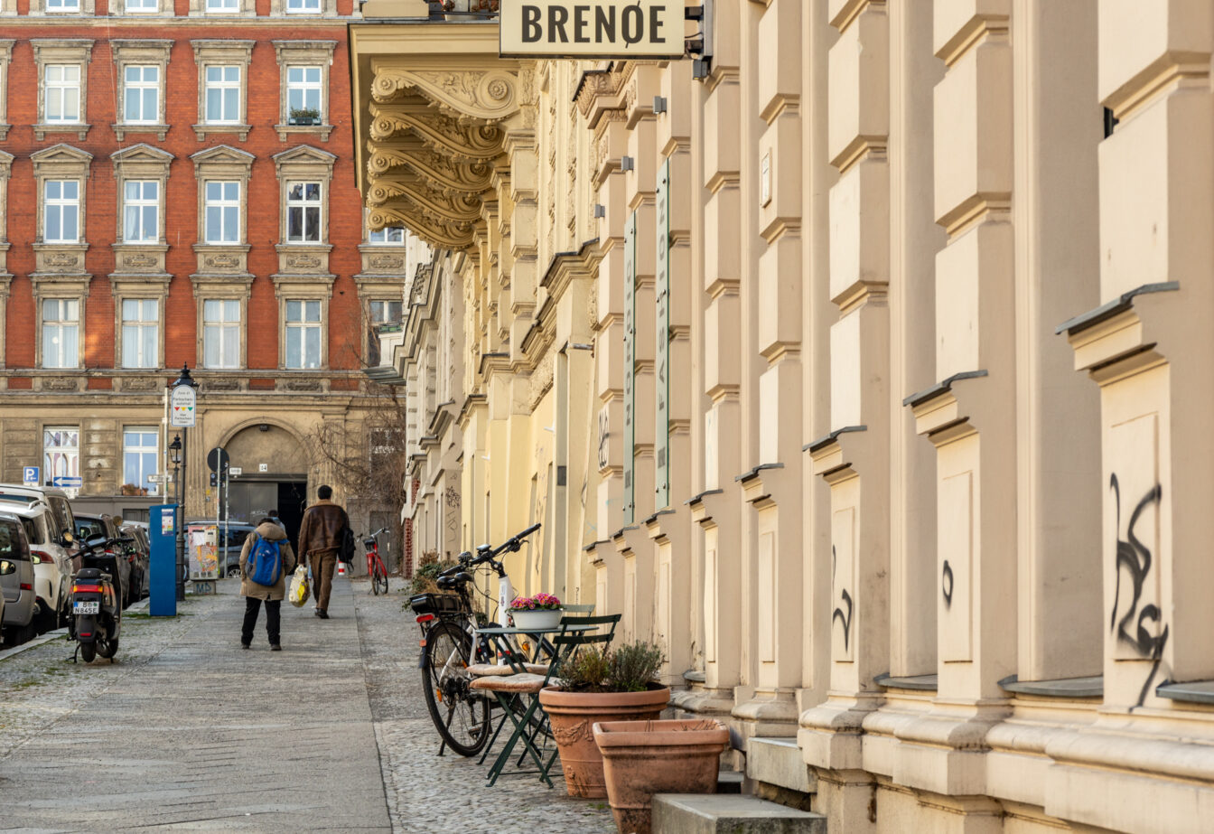 Ein belebter Straßenabschnitt im Bergmannkiez, einem beliebten Viertel in Berlin-Kreuzberg, das für seine charmante Altbauarchitektur und lebendige Atmosphäre bekannt ist. Im Vordergrund sind verzierte Fassaden in Beige- und Gelbtönen mit Fenstervorsprüngen und einigen Graffiti-Markierungen zu sehen. Vor den Gebäuden stehen Fahrräder, kleine Tische mit Stühlen sowie Pflanzentöpfe, was auf die einladende Café- und Restaurantszene des Viertels hinweist. Ein Schild mit der Aufschrift „BRENQE“ hängt an einem Gebäude. Der Gehweg ist leicht ansteigend und von geparkten Autos, einem Motorroller und Laternen gesäumt. Zwei Personen gehen entlang der Straße, eine trägt einen blauen Rucksack, die andere eine Einkaufstasche. Im Hintergrund steht ein großes, rot-braunes Backsteingebäude mit verzierten Fenstern und einem offenen Torbogen. Die Szene wird von warmem Sonnenlicht erhellt, was die gemütliche und lebendige Atmosphäre des Bergmannkiezes unterstreicht.