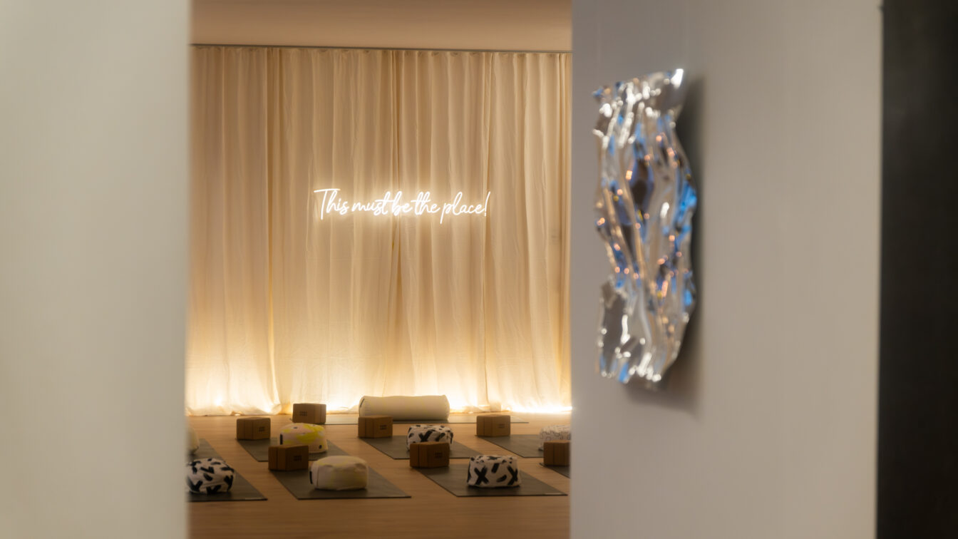 Eingang zu einem Yogastudio mit auffälliger Lichtstimmung. Am Boden liegen Matten mit Kissen. An der Wand hängt ein Leuchtschild mit der Aufschrift "This must be the place".