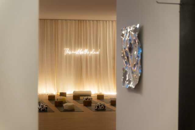 Eingang zu einem Yogastudio mit auffälliger Lichtstimmung. Am Boden liegen Matten mit Kissen. An der Wand hängt ein Leuchtschild mit der Aufschrift "This must be the place".