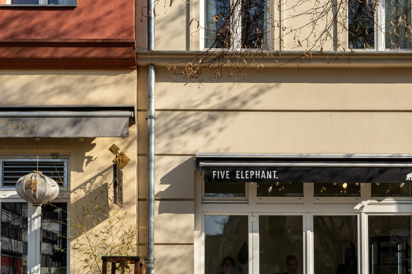 Die Fassade des beliebten Berliner Cafés Five Elephant im Stadtteil Kreuzberg. Die Außenansicht zeigt die charakteristische schwarze Markise mit weißer Schrift, die den Namen des Cafés trägt. Durch die großen Fenster lässt sich ein Blick in den Innenraum erhaschen, wo Gäste bei Kaffee und Gebäck entspannen.
Die Szene ist in warmes Sonnenlicht getaucht, wodurch sich sanfte Schatten der umliegenden Bäume auf der beigen Hauswand abzeichnen. Daneben ist ein Teil eines benachbarten Geschäfts zu sehen, das mit einem asiatisch inspirierten Lampion dekoriert ist. Die ruhige, einladende Atmosphäre spiegelt den Charme dieses Viertels wider, das für seine erstklassige Kaffeehauskultur bekannt ist. Five Elephant ist besonders berühmt für seinen exzellenten Spezialitätenkaffee und den hausgemachten New York Cheesecake, die zu den besten der Stadt zählen.