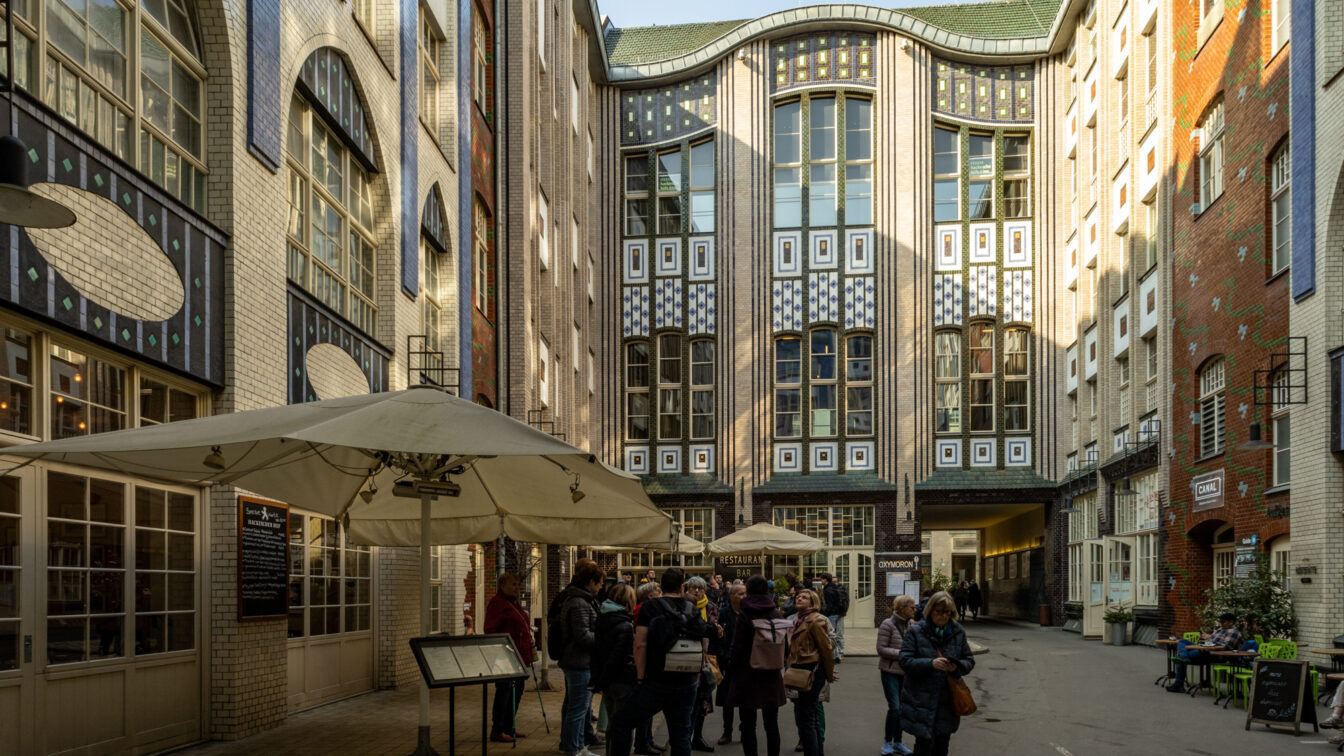 Ein belebter Innenhof in den Hackeschen Höfen in Berlin mit kunstvoll gestalteten Jugendstilfassaden aus verzierten Fliesen und großen Fenstern. Mehrere Gruppen von Menschen stehen im Hof, einige unter großen Sonnenschirmen eines Restaurants mit Außenbereich. Die Architektur zeigt eine Mischung aus weißen, blauen und braunen Kacheln mit geometrischen Mustern. Auf der rechten Seite sind Cafés und Sitzbereiche mit grünen Stühlen zu sehen, während im Hintergrund ein Durchgang zu einem weiteren Hof führt.