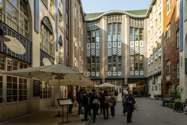 Ein belebter Innenhof in den Hackeschen Höfen in Berlin mit kunstvoll gestalteten Jugendstilfassaden aus verzierten Fliesen und großen Fenstern. Mehrere Gruppen von Menschen stehen im Hof, einige unter großen Sonnenschirmen eines Restaurants mit Außenbereich. Die Architektur zeigt eine Mischung aus weißen, blauen und braunen Kacheln mit geometrischen Mustern. Auf der rechten Seite sind Cafés und Sitzbereiche mit grünen Stühlen zu sehen, während im Hintergrund ein Durchgang zu einem weiteren Hof führt.