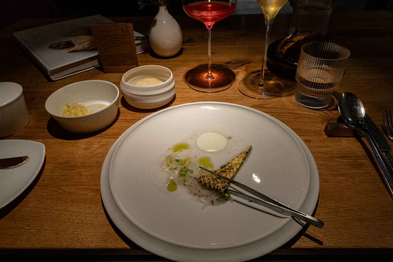 Ein kunstvoll angerichtetes Gericht im Fine-Dining-Restaurant Horváth in Berlin-Kreuzberg. Auf einem schlichten weißen Teller liegt eine filigrane Komposition aus dünn geschnittenem Fisch, einem luftigen Schaum, einem cremigen weißen Element sowie einem gegrillten Stück Gemüse, garniert mit feinen Kräutern und einigen Tropfen Öl. Eine Pinzette liegt auf dem Teller, was auf eine detailverliebte Präsentation hinweist.
Im Hintergrund auf dem elegant gedeckten Holztisch stehen feine Weingläser mit farbenfrohen Getränken, ein Wasserglas sowie kleine Schalen mit Butter und weiteren Beilagen. Ein dunkles Brot in einer schwarzen Schale deutet auf eine raffinierte Begleitung zum Menü hin. Die dezente, warme Beleuchtung verstärkt die intime Atmosphäre des Restaurants, das für seine moderne Interpretation der österreichischen Küche mit einem Fokus auf regionale Zutaten bekannt ist.