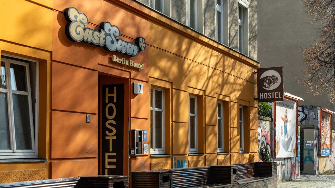 Die Fassade des 'East Seven Berlin Hostel', eines Hostels mit orangefarbenen Wänden und einem auffälligen Retro-Schild über dem Eingang. Der Schriftzug 'HOSTEL' ist groß auf der Tür zu sehen. An der Hauswand sind Schatten von Bäumen zu erkennen, die dem Gebäude eine lebendige Struktur verleihen. Rechts vom Eingang hängt ein weiteres braunes Schild mit einem Bett-Symbol und dem Wort 'HOSTEL'. Im Hintergrund sind Graffiti, Plakate und eine mit Kunst verzierte Mauer zu sehen, die auf die alternative Szene Berlins hinweisen.