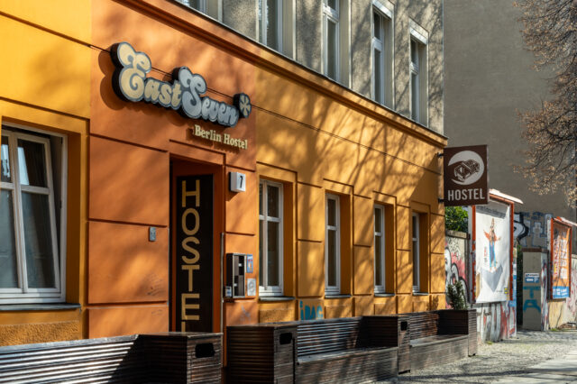 Die Fassade des 'East Seven Berlin Hostel', eines Hostels mit orangefarbenen Wänden und einem auffälligen Retro-Schild über dem Eingang. Der Schriftzug 'HOSTEL' ist groß auf der Tür zu sehen. An der Hauswand sind Schatten von Bäumen zu erkennen, die dem Gebäude eine lebendige Struktur verleihen. Rechts vom Eingang hängt ein weiteres braunes Schild mit einem Bett-Symbol und dem Wort 'HOSTEL'. Im Hintergrund sind Graffiti, Plakate und eine mit Kunst verzierte Mauer zu sehen, die auf die alternative Szene Berlins hinweisen.