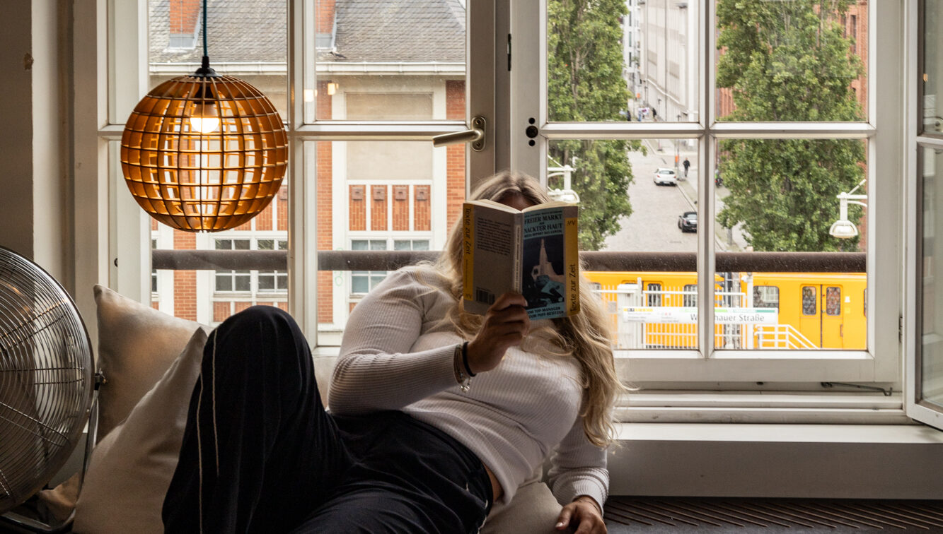 Szene aus einem Zimmer des Michelberger Hotels in Berlin: Eine Frau liegt entspannt auf einer Holzplattform am Fenster, liest ein Buch und blickt dabei ins Freie. Hinter ihr ein großes, geöffnetes Sprossenfenster mit Blick auf eine ruhige Berliner Straße, hohe Bäume und eine vorbeifahrende gelbe Straßenbahn. Links im Bild eine hängende, kugelförmige Lampe aus Holzstreben und ein Ventilator auf einem Kissen. Die Atmosphäre ist gemütlich, urban und entspannt.