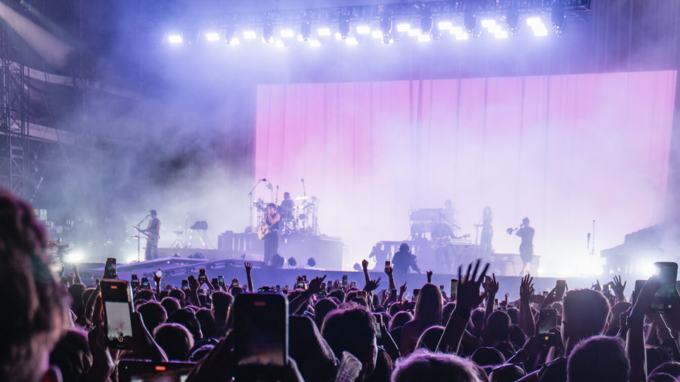 Große Menschenmenge bei einem Open-Air-Festival in Europa, viele halten ihre Smartphones hoch. Auf der Bühne spielen mehrere Musiker vor einer großen, rosa beleuchteten Leinwand, umgeben von Nebel und starken Scheinwerfern.