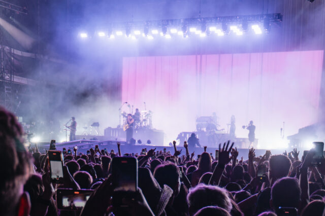 Große Menschenmenge bei einem Open-Air-Festival in Europa, viele halten ihre Smartphones hoch. Auf der Bühne spielen mehrere Musiker vor einer großen, rosa beleuchteten Leinwand, umgeben von Nebel und starken Scheinwerfern.