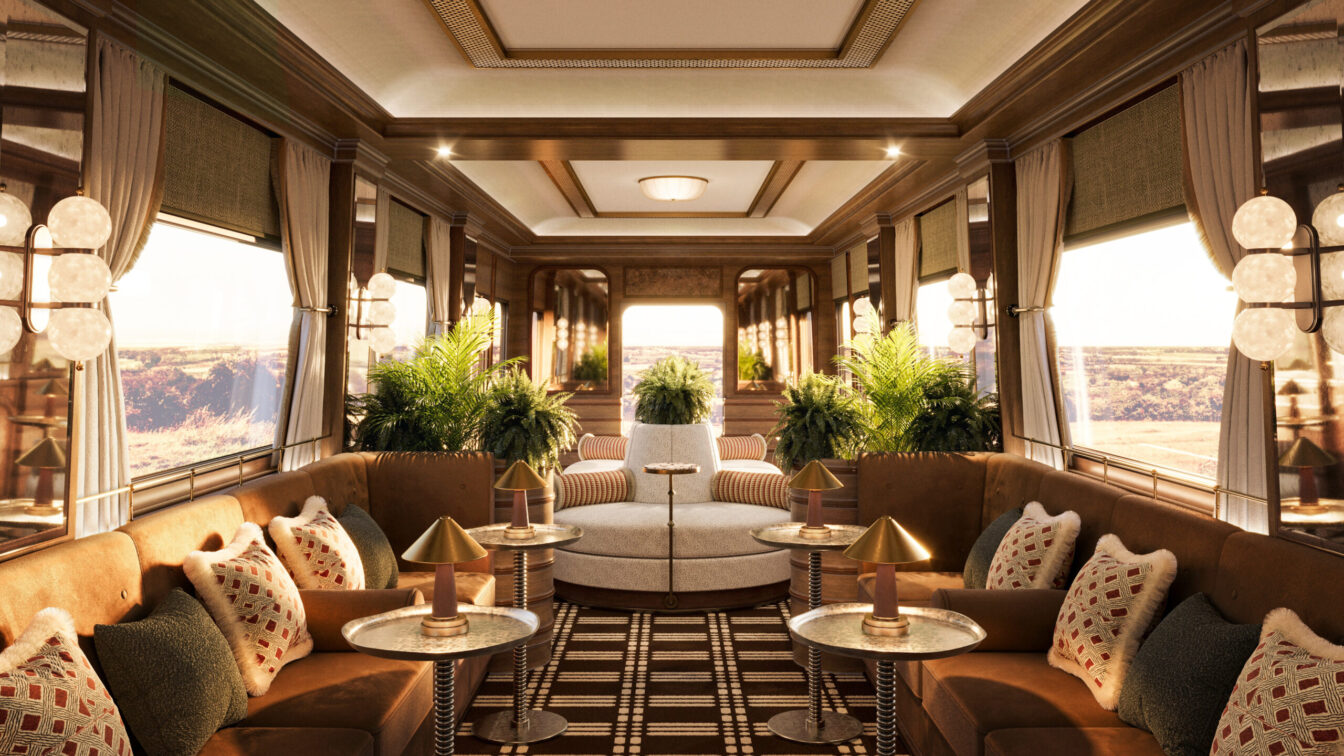 Luxuriöses Interieur eines historischen Zugwaggons mit edler Holzverkleidung, großen Fenstern und stilvoller Beleuchtung. Die Lounge ist mit samtigen Polstern in warmen Brauntönen, dekorativen Kissen und eleganten Beistelltischen mit goldenen Lampen ausgestattet. Üppige grüne Pflanzen verleihen dem Raum eine frische Note, während ein geometrisch gemusterter Teppich den Boden schmückt. Die Fenster geben den Blick auf eine malerische Landschaft frei, die in warmes Sonnenlicht getaucht ist.