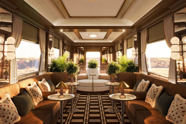 Luxuriöses Interieur eines historischen Zugwaggons mit edler Holzverkleidung, großen Fenstern und stilvoller Beleuchtung. Die Lounge ist mit samtigen Polstern in warmen Brauntönen, dekorativen Kissen und eleganten Beistelltischen mit goldenen Lampen ausgestattet. Üppige grüne Pflanzen verleihen dem Raum eine frische Note, während ein geometrisch gemusterter Teppich den Boden schmückt. Die Fenster geben den Blick auf eine malerische Landschaft frei, die in warmes Sonnenlicht getaucht ist.