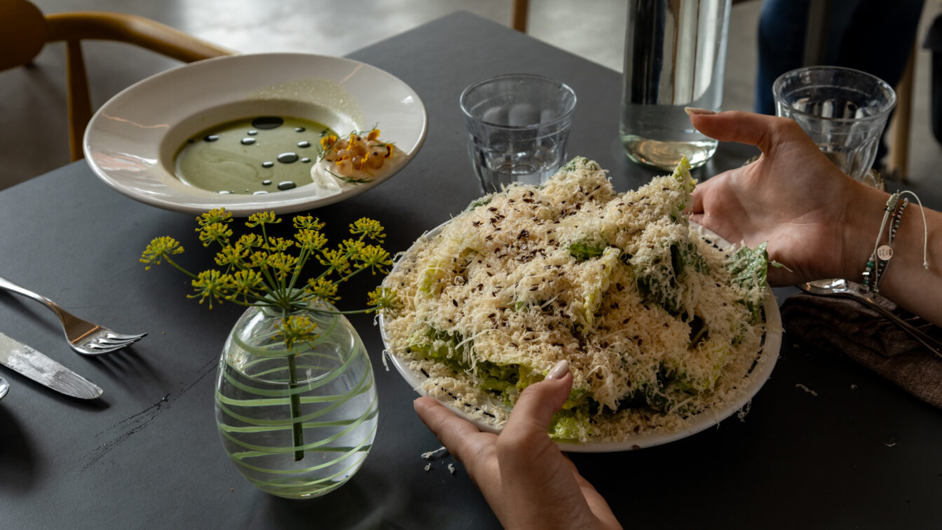 Detailaufnahme eines kunstvoll angerichteten Essens in einem außergewöhnlichen Restaurant in Berlin: Ein großer Caesar Salad mit reichlich frisch geriebenem Käse und eine grüne Suppe mit Öl-Perlen, serviert in modernem Ambiente mit stilvoller Tischdekoration – ein kulinarisches Erlebnis für Feinschmecker.
