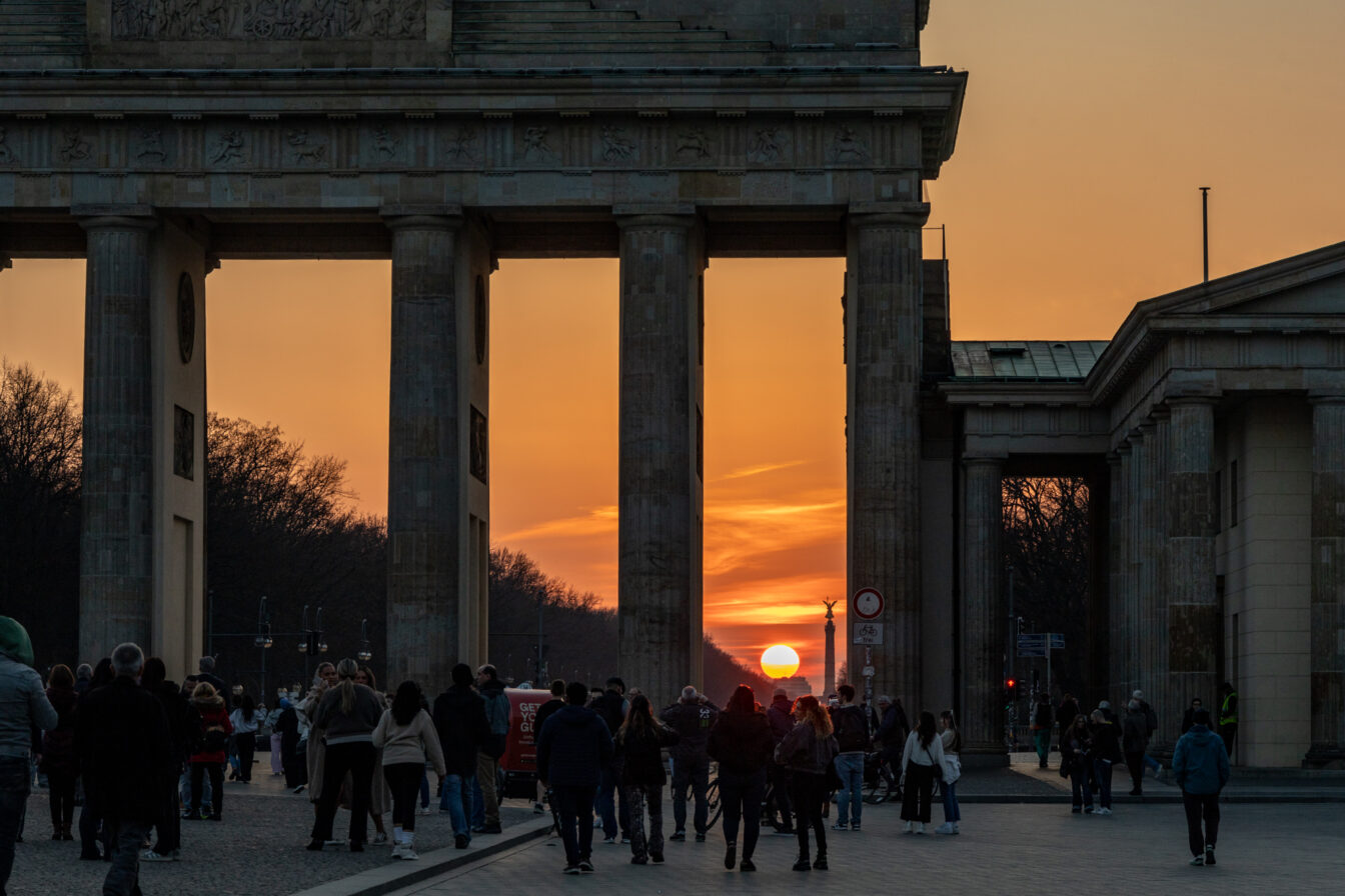 Menschen spazieren bei Sonnenuntergang durch das Brandenburger Tor in Berlin. Die Sonne steht tief am Horizont und scheint genau durch die Säulen des Tores. Der Himmel ist orange gefärbt, Silhouetten der Menschen und Gebäude rahmen die Szene stimmungsvoll ein.