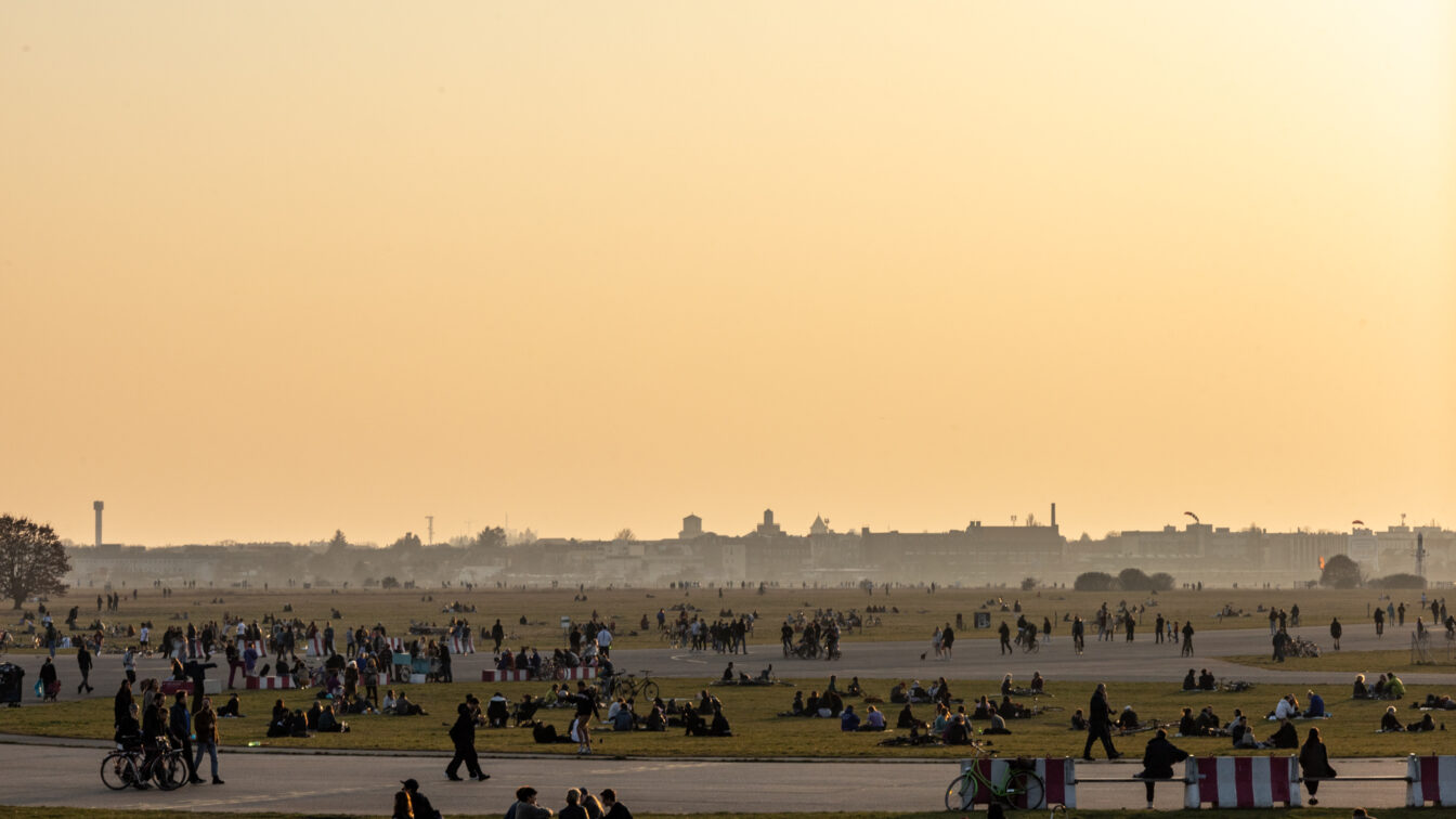 Menschen genießen einen warmen Sommertag im Tempelhofer Feld in Berlin. Der weitläufige ehemalige Flughafen dient als Park, wo zahlreiche Menschen auf der Wiese sitzen, spazieren gehen, Fahrrad fahren oder in Gruppen zusammenkommen. Im Hintergrund ist die Skyline Berlins mit historischen und modernen Gebäuden zu sehen, während der Himmel in warmen orangefarbenen Tönen der Abenddämmerung leuchtet.