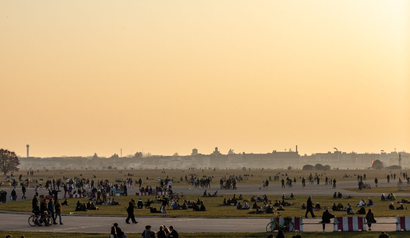 Menschen genießen einen warmen Sommertag im Tempelhofer Feld in Berlin. Der weitläufige ehemalige Flughafen dient als Park, wo zahlreiche Menschen auf der Wiese sitzen, spazieren gehen, Fahrrad fahren oder in Gruppen zusammenkommen. Im Hintergrund ist die Skyline Berlins mit historischen und modernen Gebäuden zu sehen, während der Himmel in warmen orangefarbenen Tönen der Abenddämmerung leuchtet.