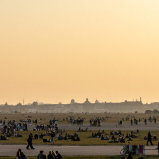 Menschen genießen einen warmen Sommertag im Tempelhofer Feld in Berlin. Der weitläufige ehemalige Flughafen dient als Park, wo zahlreiche Menschen auf der Wiese sitzen, spazieren gehen, Fahrrad fahren oder in Gruppen zusammenkommen. Im Hintergrund ist die Skyline Berlins mit historischen und modernen Gebäuden zu sehen, während der Himmel in warmen orangefarbenen Tönen der Abenddämmerung leuchtet.
