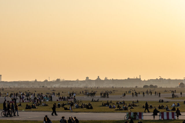 Menschen genießen einen warmen Sommertag im Tempelhofer Feld in Berlin. Der weitläufige ehemalige Flughafen dient als Park, wo zahlreiche Menschen auf der Wiese sitzen, spazieren gehen, Fahrrad fahren oder in Gruppen zusammenkommen. Im Hintergrund ist die Skyline Berlins mit historischen und modernen Gebäuden zu sehen, während der Himmel in warmen orangefarbenen Tönen der Abenddämmerung leuchtet.