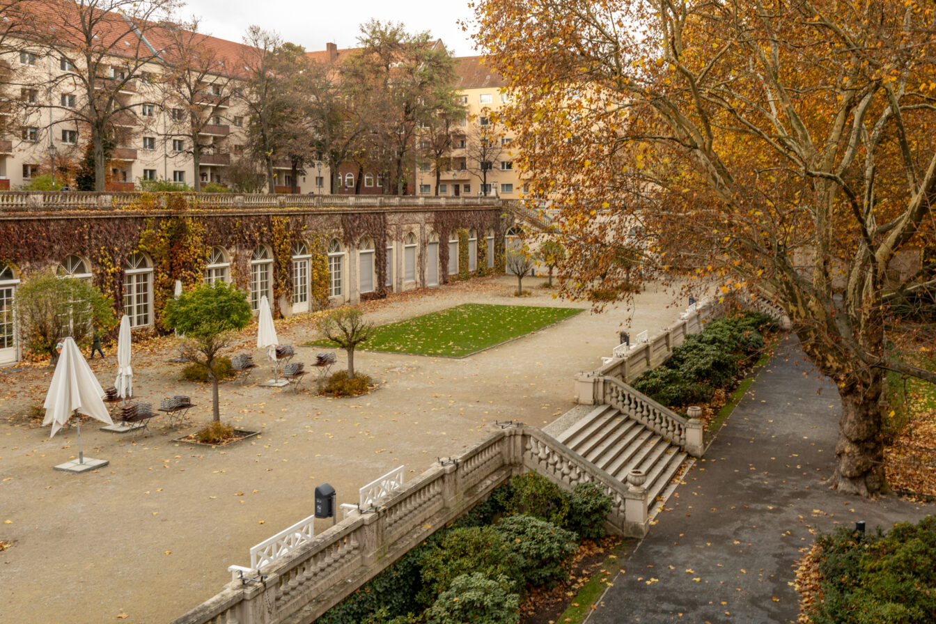Ein herbstlicher Blick auf den Körnerpark in Berlin. Die historische Parkanlage ist von einer niedrigen Balustrade umgeben, mit einer breiten Treppe, die hinunterführt. Die Bäume tragen golden verfärbte Blätter, und einige haben bereits ihr Laub verloren. In der Mitte des Parks befindet sich eine gepflegte Rasenfläche, umgeben von Schotterwegen mit Sitzgelegenheiten und geschlossenen Sonnenschirmen. Die historische Orangerie mit ihren bogenförmigen Fenstern und mit Efeu bewachsenen Fassaden bildet den Hintergrund, während dahinter Wohnhäuser mit roten Dächern zu sehen sind.