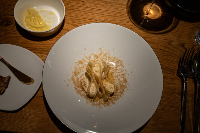 Ein kunstvoll angerichtetes Pastagericht auf einem weißen Teller: Die Pasta ist elegant gefaltet und mit einer cremigen Sauce überzogen, umgeben von feinen, geriebenen Zutaten. Die Szene ist in warmem Licht gehalten, mit einem Glas Wein und weiteren Begleitkomponenten auf einem rustikalen Holztisch.