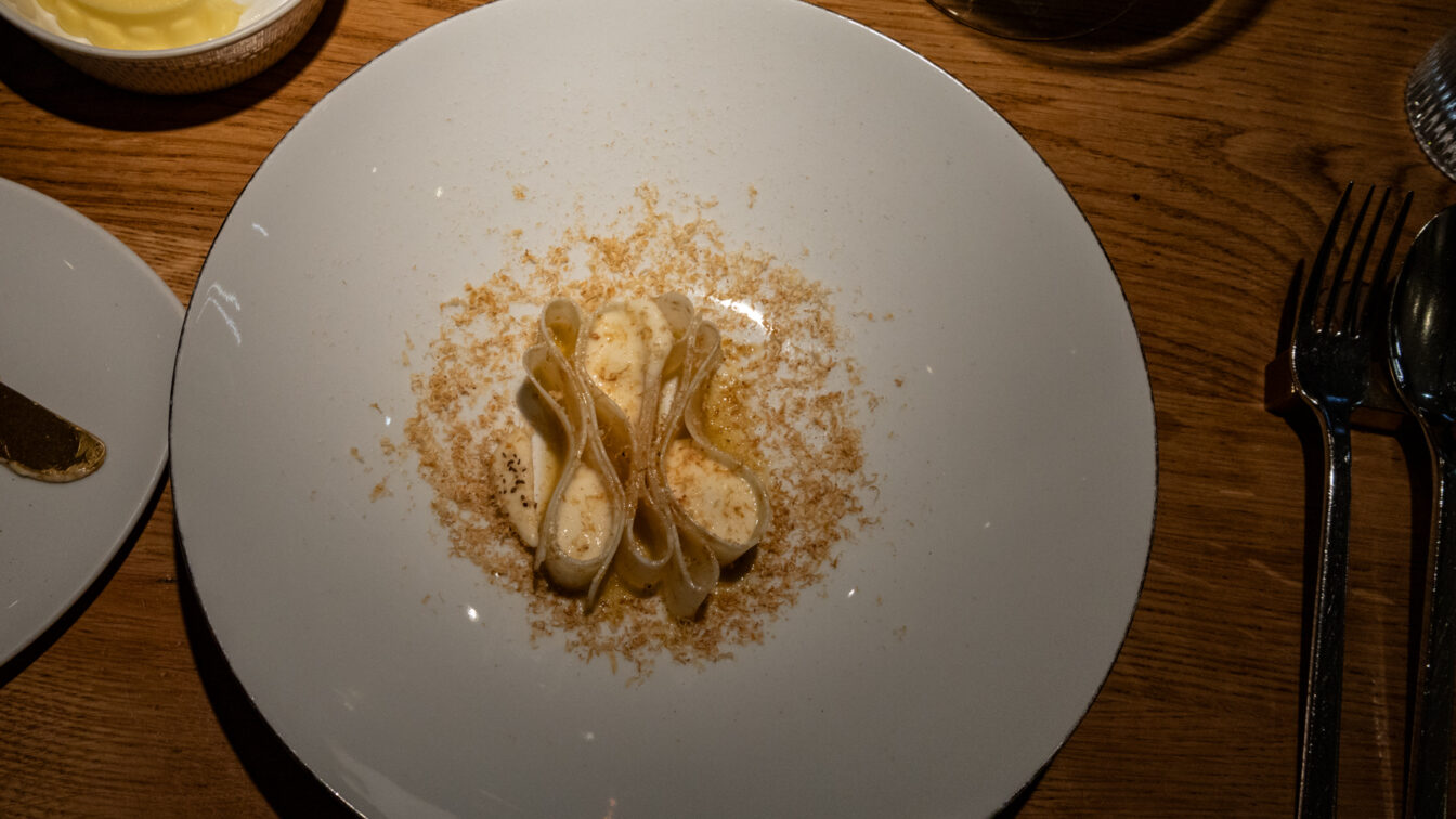 Ein kunstvoll angerichtetes Pastagericht auf einem weißen Teller: Die Pasta ist elegant gefaltet und mit einer cremigen Sauce überzogen, umgeben von feinen, geriebenen Zutaten. Die Szene ist in warmem Licht gehalten, mit einem Glas Wein und weiteren Begleitkomponenten auf einem rustikalen Holztisch.