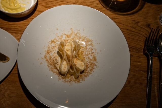 Ein kunstvoll angerichtetes Pastagericht auf einem weißen Teller: Die Pasta ist elegant gefaltet und mit einer cremigen Sauce überzogen, umgeben von feinen, geriebenen Zutaten. Die Szene ist in warmem Licht gehalten, mit einem Glas Wein und weiteren Begleitkomponenten auf einem rustikalen Holztisch.