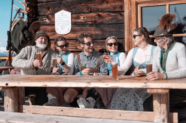 Personen in Tracht sitzen vor der Hütte und stoßen gutgelaunt an