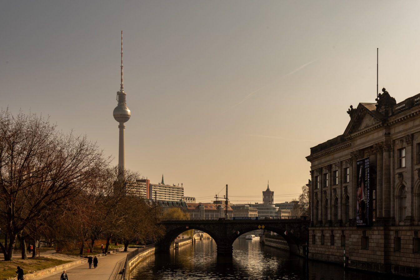 Blick vom Monbijoupark in Berlin auf den Fernsehturm bei Sonnenuntergang. Im Vordergrund verläuft die Spree mit einer Brücke darüber, auf der Züge fahren. Rechts das Bode-Museum mit klassizistischer Fassade und einem großen Banner. Links der Uferweg mit Spaziergängern und kahlen Bäumen im Winter oder frühen Frühling. Die Szene ist in warmes, goldenes Licht getaucht und vermittelt eine ruhige, stimmungsvolle Atmosphäre.