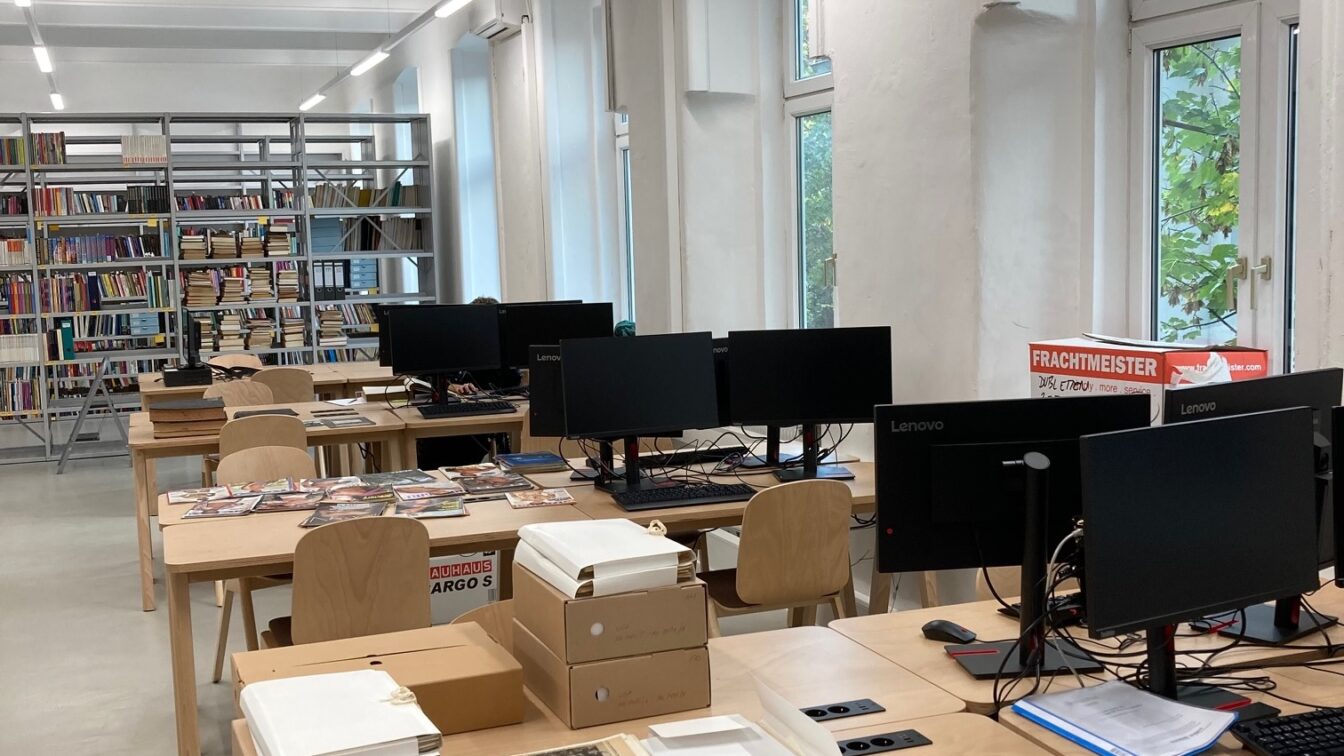 Das Bild zeigt einen modernen Raum, der wie ein Arbeitsbereich in einer Bibliothek oder einem Archiv aussieht. Es gibt mehrere Tische mit Computern und Büchern. Auf den Tischen liegen Zeitschriften und einige Dokumente, die teilweise in weiße Ordner verpackt sind. Im Hintergrund sind Regale mit einer Vielzahl von Büchern sichtbar. Große Fenster lassen natürliches Licht in den Raum, und im hinteren Bereich des Raums ist ein Kasten mit Aufschrift "Frachtmeister" zu sehen. Ein Mitarbeiter ist an einem der Computer zu sehen, während er an einem Bildschirm arbeitet.