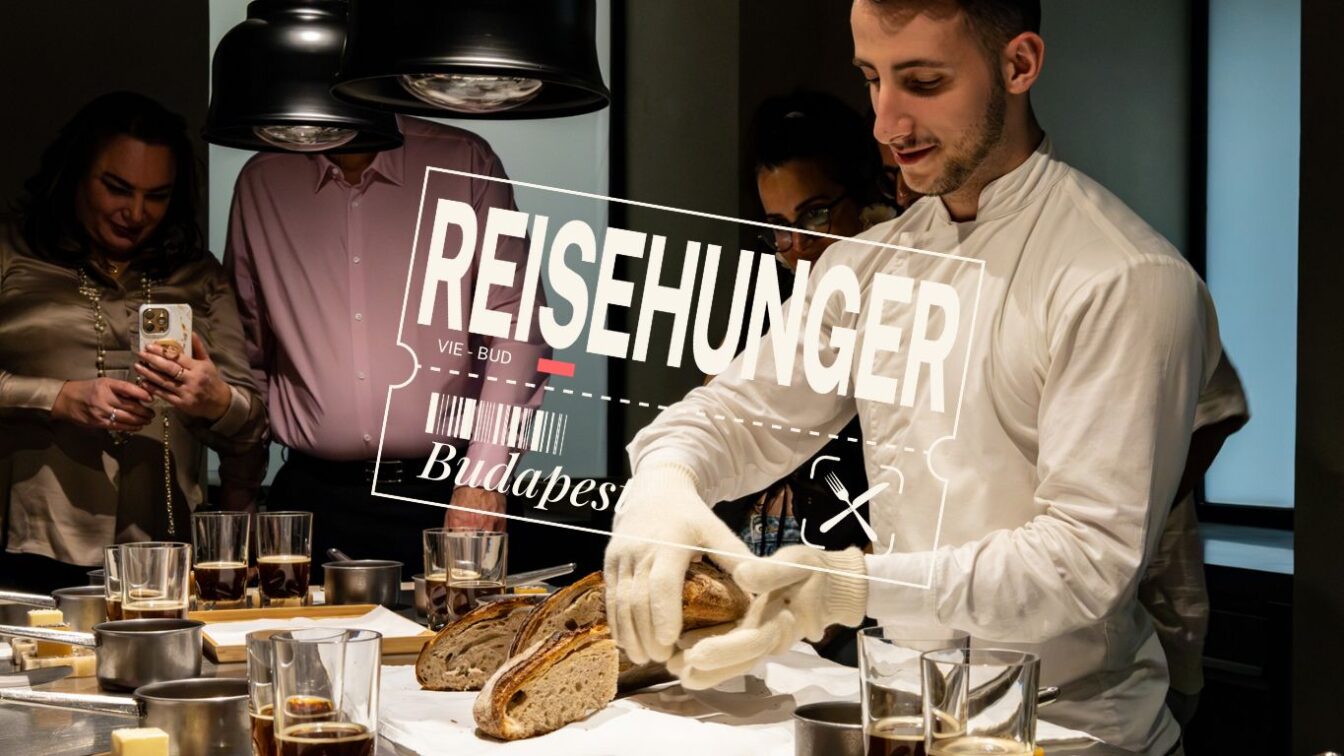 Ein junger Koch in weißer Kochjacke und Handschuhen schneidet frisches Sauerteigbrot bei einem Tasting im Onyx Műhely in Budapest. Um ihn herum stehen mehrere Gäste, die das Erlebnis fotografieren und beobachten. Auf dem Tisch befinden sich Gläser mit Getränken und kleine Töpfe. Im Vordergrund ist der Schriftzug „REISEHUNGER – VIE-BUD – Budapest“ eingeblendet.