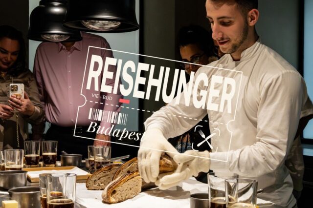 Ein junger Koch in weißer Kochjacke und Handschuhen schneidet frisches Sauerteigbrot bei einem Tasting im Onyx Műhely in Budapest. Um ihn herum stehen mehrere Gäste, die das Erlebnis fotografieren und beobachten. Auf dem Tisch befinden sich Gläser mit Getränken und kleine Töpfe. Im Vordergrund ist der Schriftzug „REISEHUNGER – VIE-BUD – Budapest“ eingeblendet.