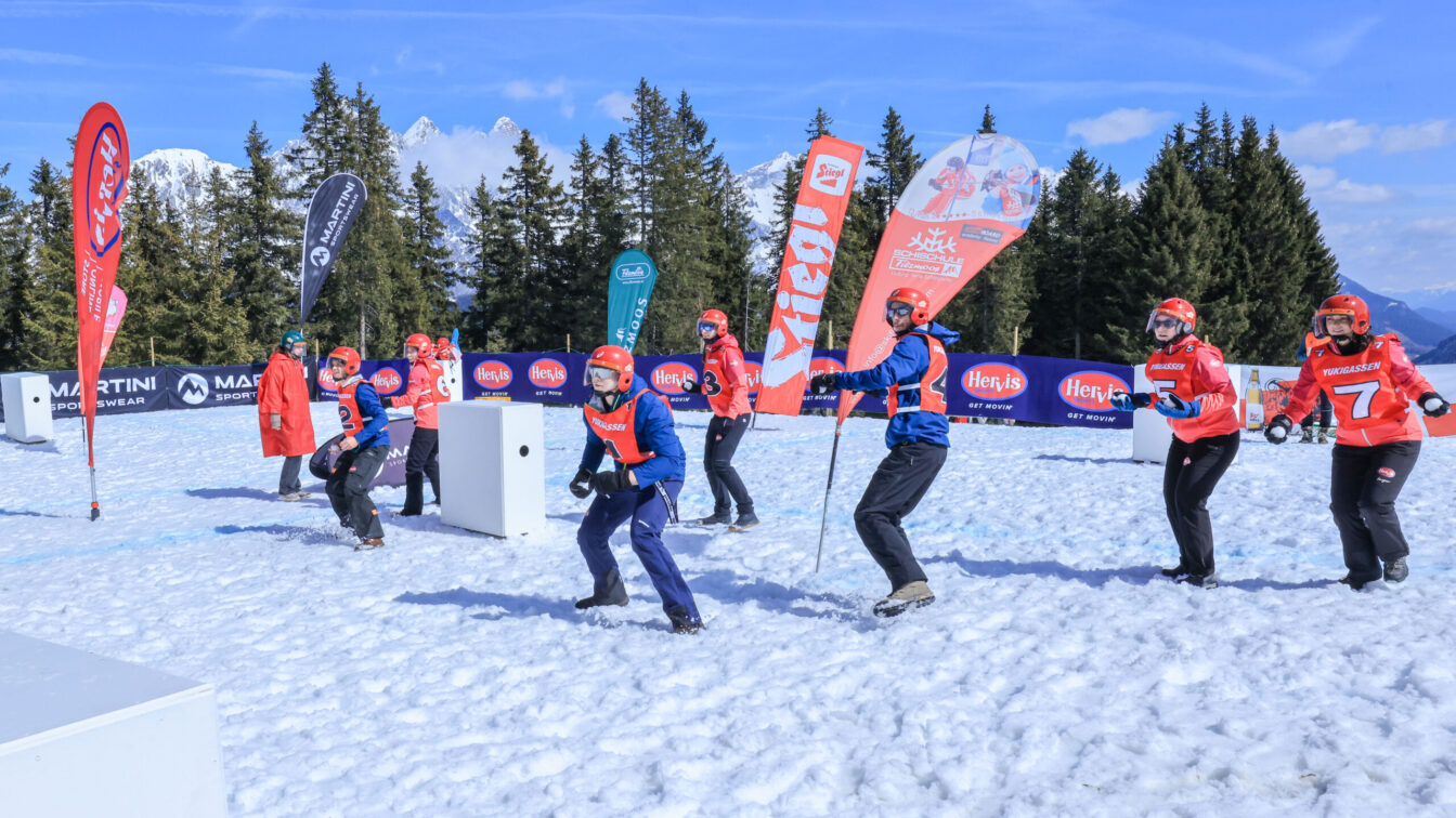 Eine Gruppe von Menschen nimmt an einer Schneeballschlacht-Wettbewerb teil. Sie tragen Helme, Schutzbrillen und farbige Trikots mit Nummern. Die Szene spielt sich auf einer schneebedeckten Fläche in den Bergen ab, mit blauen Bannern und bunten Fahnen im Hintergrund. Die Teilnehmer bewegen sich aktiv, einige in Abwehrhaltung, andere scheinbar zum Wurf bereit. Die Sonne scheint, und der Himmel ist klar mit wenigen Wolken. Im Hintergrund sind Nadelbäume und schneebedeckte Berggipfel zu sehen.