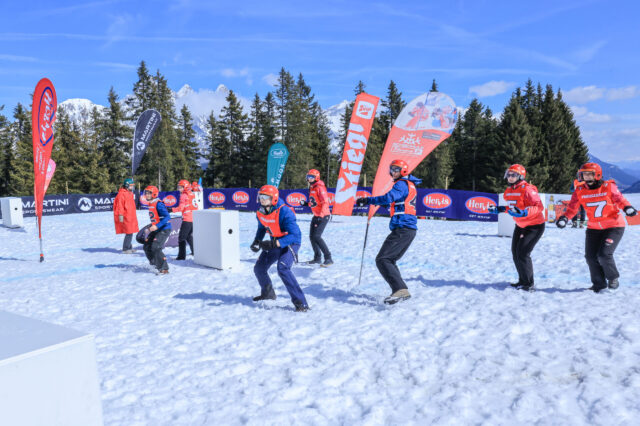Eine Gruppe von Menschen nimmt an einer Schneeballschlacht-Wettbewerb teil. Sie tragen Helme, Schutzbrillen und farbige Trikots mit Nummern. Die Szene spielt sich auf einer schneebedeckten Fläche in den Bergen ab, mit blauen Bannern und bunten Fahnen im Hintergrund. Die Teilnehmer bewegen sich aktiv, einige in Abwehrhaltung, andere scheinbar zum Wurf bereit. Die Sonne scheint, und der Himmel ist klar mit wenigen Wolken. Im Hintergrund sind Nadelbäume und schneebedeckte Berggipfel zu sehen.