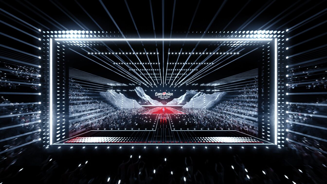 Das Bild zeigt eine beeindruckende Szene vom Eurovision Song Contest 2025 in Basel, mit einer modernen und futuristischen Bühne. Die Bühne ist von hellen Lichtstrahlen umgeben, die dynamisch durch den Raum verlaufen und den Fokus auf das große Event lenken. Auf der Bühne ist das "Eurovision Song Contest"-Logo prominent zu sehen. Im Hintergrund ist das Publikum mit vielen Handylichtern zu sehen, die eine festliche Atmosphäre schaffen. Die Beleuchtung und das Design der Bühne unterstreichen das spektakuläre und energiegeladene Ambiente dieses internationalen Musikwettbewerbs.