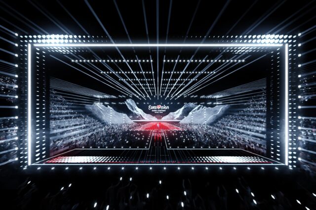 Das Bild zeigt eine beeindruckende Szene vom Eurovision Song Contest 2025 in Basel, mit einer modernen und futuristischen Bühne. Die Bühne ist von hellen Lichtstrahlen umgeben, die dynamisch durch den Raum verlaufen und den Fokus auf das große Event lenken. Auf der Bühne ist das "Eurovision Song Contest"-Logo prominent zu sehen. Im Hintergrund ist das Publikum mit vielen Handylichtern zu sehen, die eine festliche Atmosphäre schaffen. Die Beleuchtung und das Design der Bühne unterstreichen das spektakuläre und energiegeladene Ambiente dieses internationalen Musikwettbewerbs.