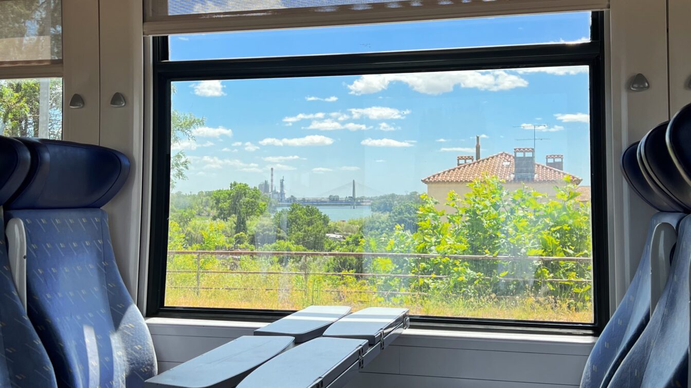 Zugfahrt Frankreich Barcelona Italien, Blick aus dem Fenster eines Zugabteils mit blauen Sitzen und Klapptischen. Draußen ist eine grüne Landschaft mit Bäumen, einem Haus mit rotem Ziegeldach und einer Brücke im Hintergrund zu sehen. Der Himmel ist blau mit einigen weißen Wolken.