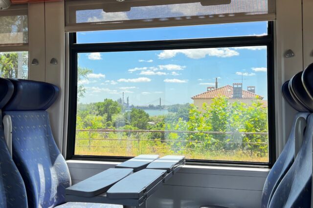 Zugfahrt Frankreich Barcelona Italien, Blick aus dem Fenster eines Zugabteils mit blauen Sitzen und Klapptischen. Draußen ist eine grüne Landschaft mit Bäumen, einem Haus mit rotem Ziegeldach und einer Brücke im Hintergrund zu sehen. Der Himmel ist blau mit einigen weißen Wolken.