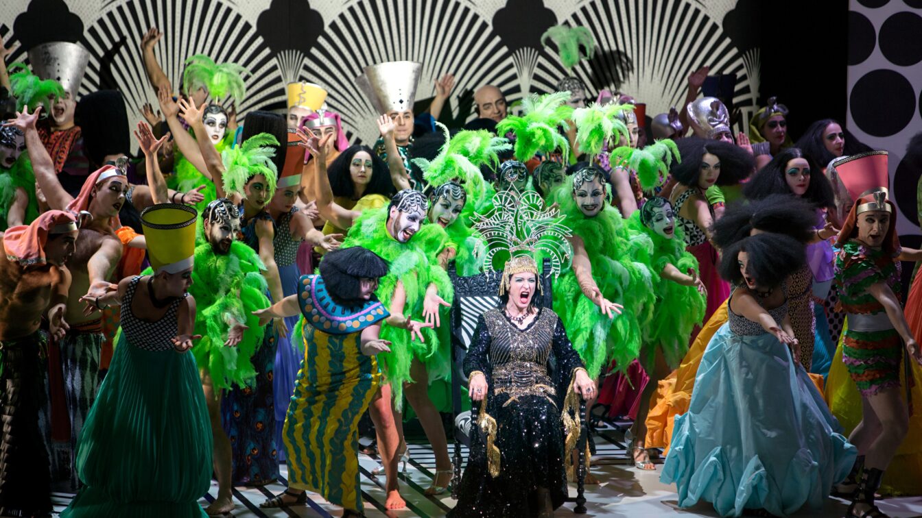 Schillernde Bühnensituation mit vielen Personen in bunten Kostümen mit grünen Federn und auffällig geschminkten Gesichtern