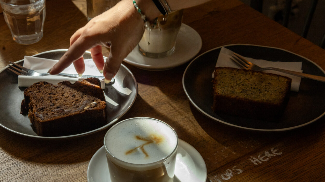 Gemütliche Café-Szene in Berlin: Auf einem Holztisch stehen zwei Stücke Kuchen auf dunklen Tellern, Cappuccino mit Latte Art und ein Glas Wasser. Eine Hand greift nach Besteck im warmen Licht – typisches Berliner Café-Flair mit entspannter Atmosphäre und hausgemachtem Gebäck.