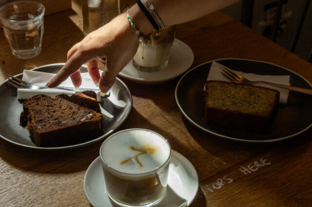 Gemütliche Café-Szene in Berlin: Auf einem Holztisch stehen zwei Stücke Kuchen auf dunklen Tellern, Cappuccino mit Latte Art und ein Glas Wasser. Eine Hand greift nach Besteck im warmen Licht – typisches Berliner Café-Flair mit entspannter Atmosphäre und hausgemachtem Gebäck.