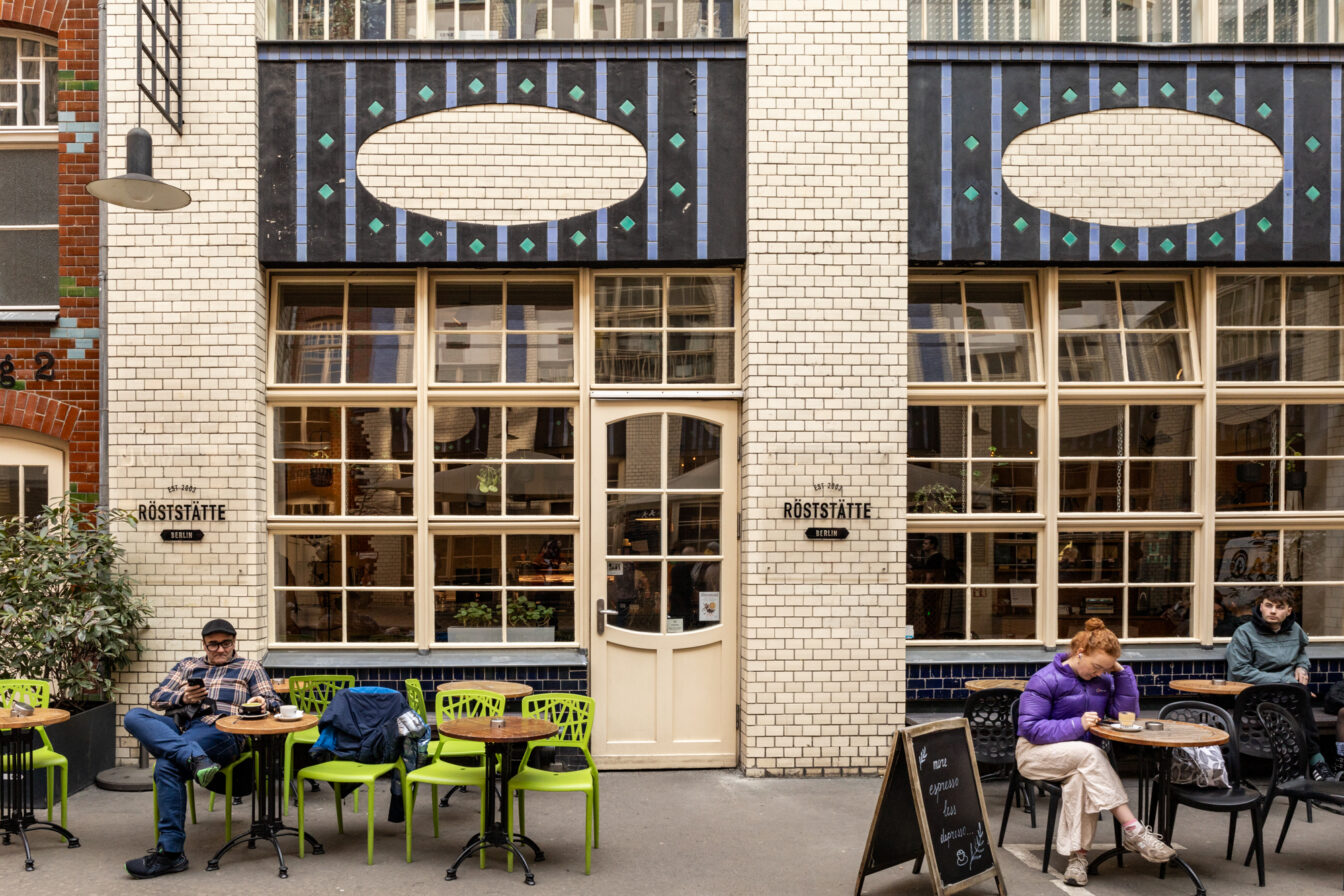Außenansicht des Cafés „Röststätte Berlin“ in einem charmanten Hinterhof mit heller Klinkerfassade und auffälligen Verzierungen. Vor dem Café sitzen mehrere Menschen an kleinen Tischen, genießen Kaffee und wirken entspannt. Eine Tafel bewirbt Espresso. Die Atmosphäre ist ruhig und urban.