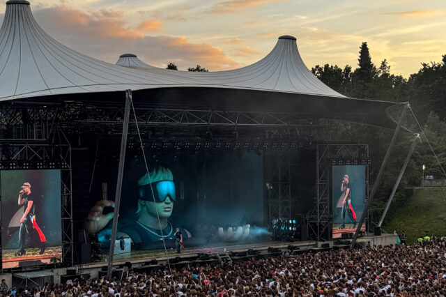 Großes Open-Air-Konzert im Sommer 2025 in Berlin: Eine riesige Menschenmenge steht vor der überdachten Bühne der Waldbühne. Auf der Bühne ein Künstler mit Sonnenbrille, unterstützt von beeindruckenden LED-Effekten und einer riesigen futuristischen Bühnenfigur. Der Himmel zeigt ein farbenfrohes Abendlicht – perfekte Festivalstimmung in Berlin.