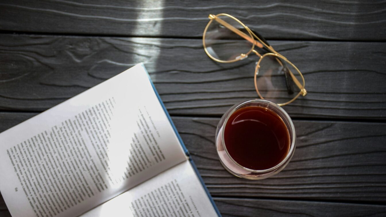 Ein aufgeschlagenes Buch neben einem Glas Rotwein und einer Lesebrille.