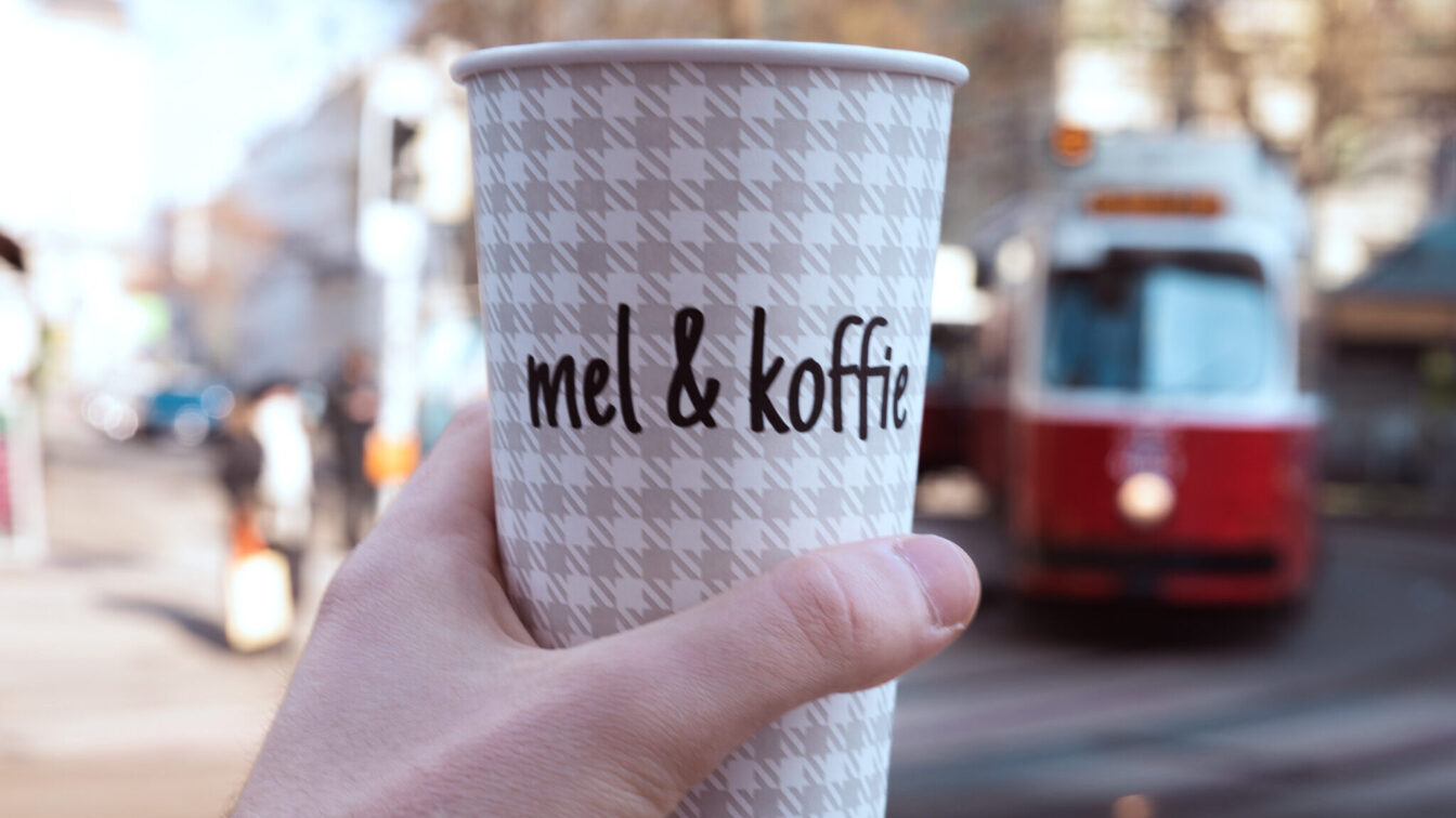 Ein Kaffeebecher von Mel&koffie, im Hintergrund ist verschwommen eine rote Wiener Straßenbahn zu sehen
