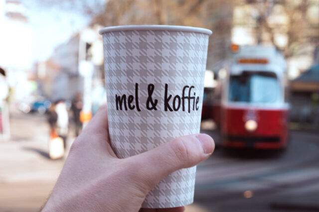 Ein Kaffeebecher von Mel&koffie, im Hintergrund ist verschwommen eine rote Wiener Straßenbahn zu sehen