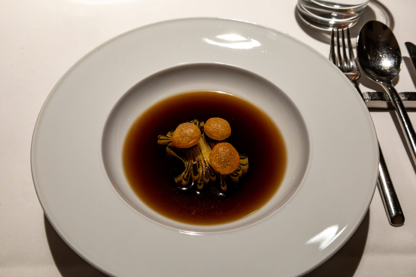Ein eleganter, weißer Teller mit tiefer Mulde, darin eine klare, dunkelbraune Brühe. In der Mitte eine kunstvolle Gemüsekomposition, vermutlich geschmorter Lauch oder Sellerie, vertikal angerichtet und mit drei kleinen goldbraunen Chips garniert. Das minimalistische Setting mit glänzendem Besteck und weißer Tischdecke unterstreicht den Fine-Dining-Charakter des Gerichts.