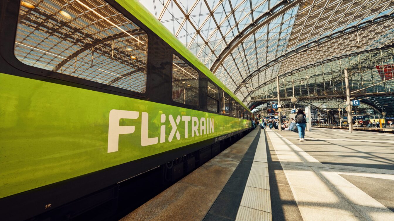 Ein grüner FlixTrain steht an einem Bahnsteig in einem modernen, lichtdurchfluteten Bahnhof mit gläsernem Dach. Menschen laufen entlang des Bahnsteigs, einige mit Gepäck. Die Perspektive zeigt den Zug in Nahaufnahme auf der linken Seite, während sich das Glasdach bogenförmig über die gesamte Szene spannt.